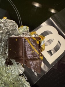Chocolate bar wrapped in silver foil and partly unwrapped, next to packaging with white and black logo. Yellow and purple designs on the chocolate.