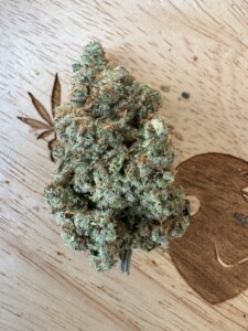 Cannabis bud with green and orange trichomes on a wood grain surface; a leaf and a brown circle are also present.