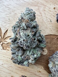 A cannabis bud with frosty trichomes, on a wooden surface with a cannabis leaf design.