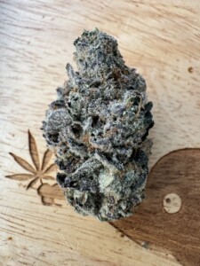 A single cannabis bud with frosted trichomes on a wooden surface, with a carved leaf and a wooden decorative element.