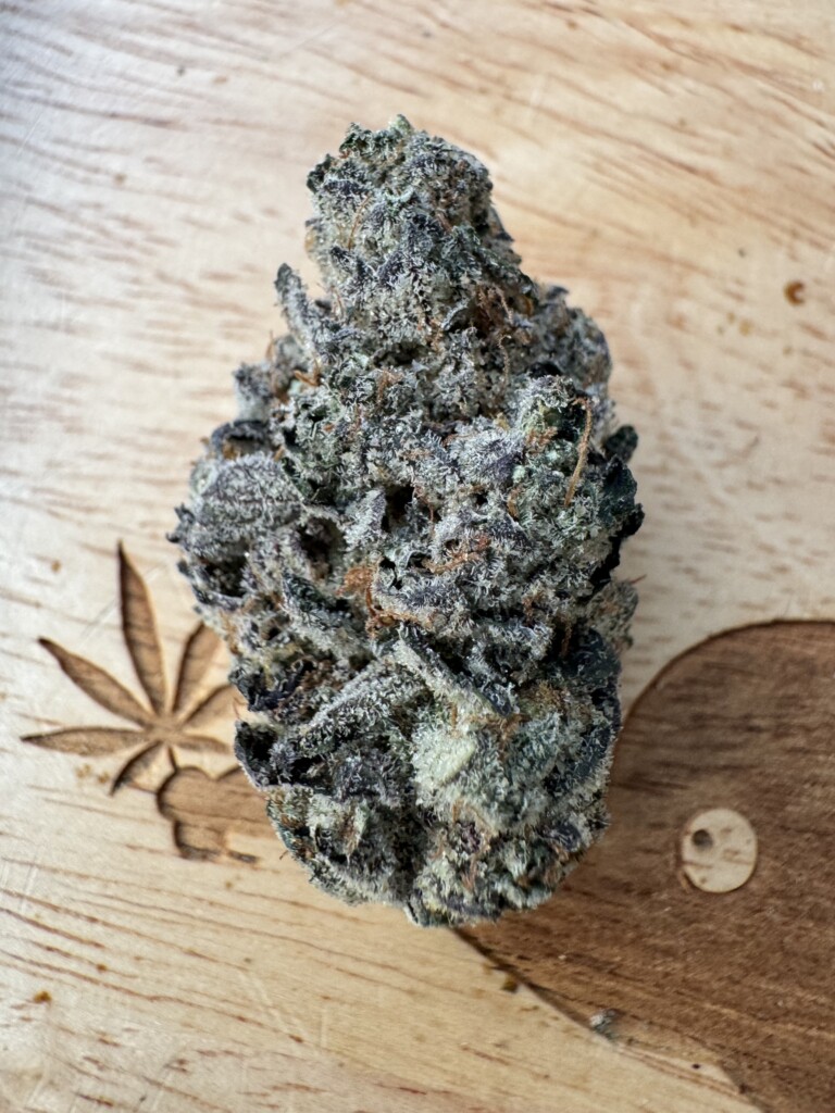 A single cannabis bud with frosted trichomes on a wooden surface, with a carved leaf and a wooden decorative element.