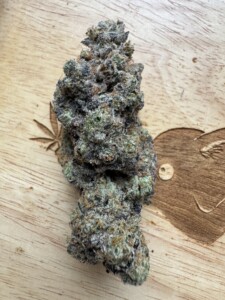 A large cannabis bud with dense trichomes and visible purple and green hues, on a wooden surface with carved leaf designs.