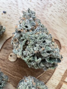 Cannabis bud with green and brown hues, covered in white trichomes, on a wooden surface.